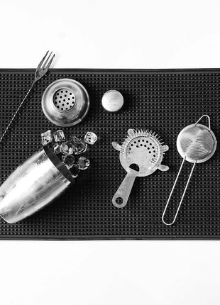 Bar tools including a jigger, strainer, and shaker on a black bar mat.