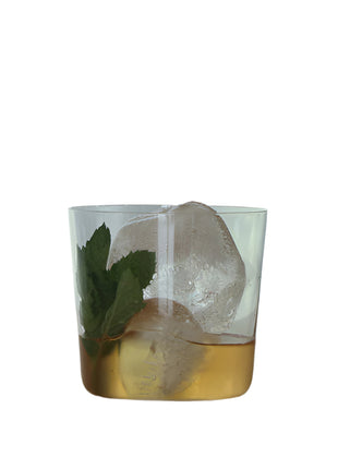 Glass of amber liquid with a large ice cube and mint leaf on a reflective surface.